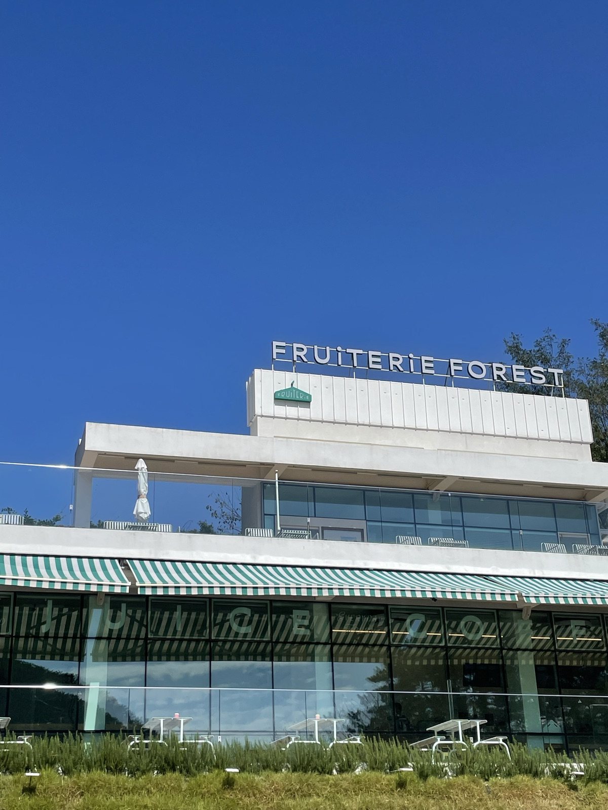 파란 하늘 아래 FRUITERIE FOREST 간판이 달린 흰 건물과 초록 줄무늬 차양, 유리창 앞 야외 테이블