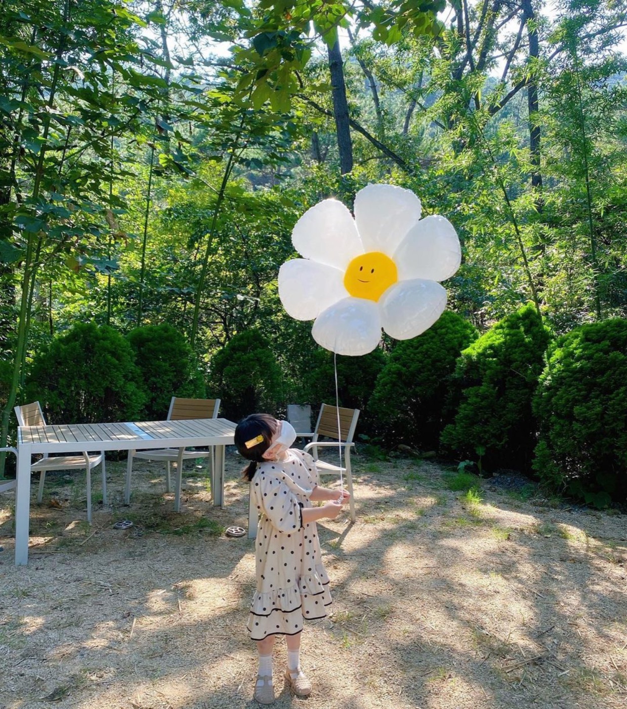 숲속 정원에서 흰 꽃 모양 풍선을 든 아이와 나무 그늘 아래 야외 테이블